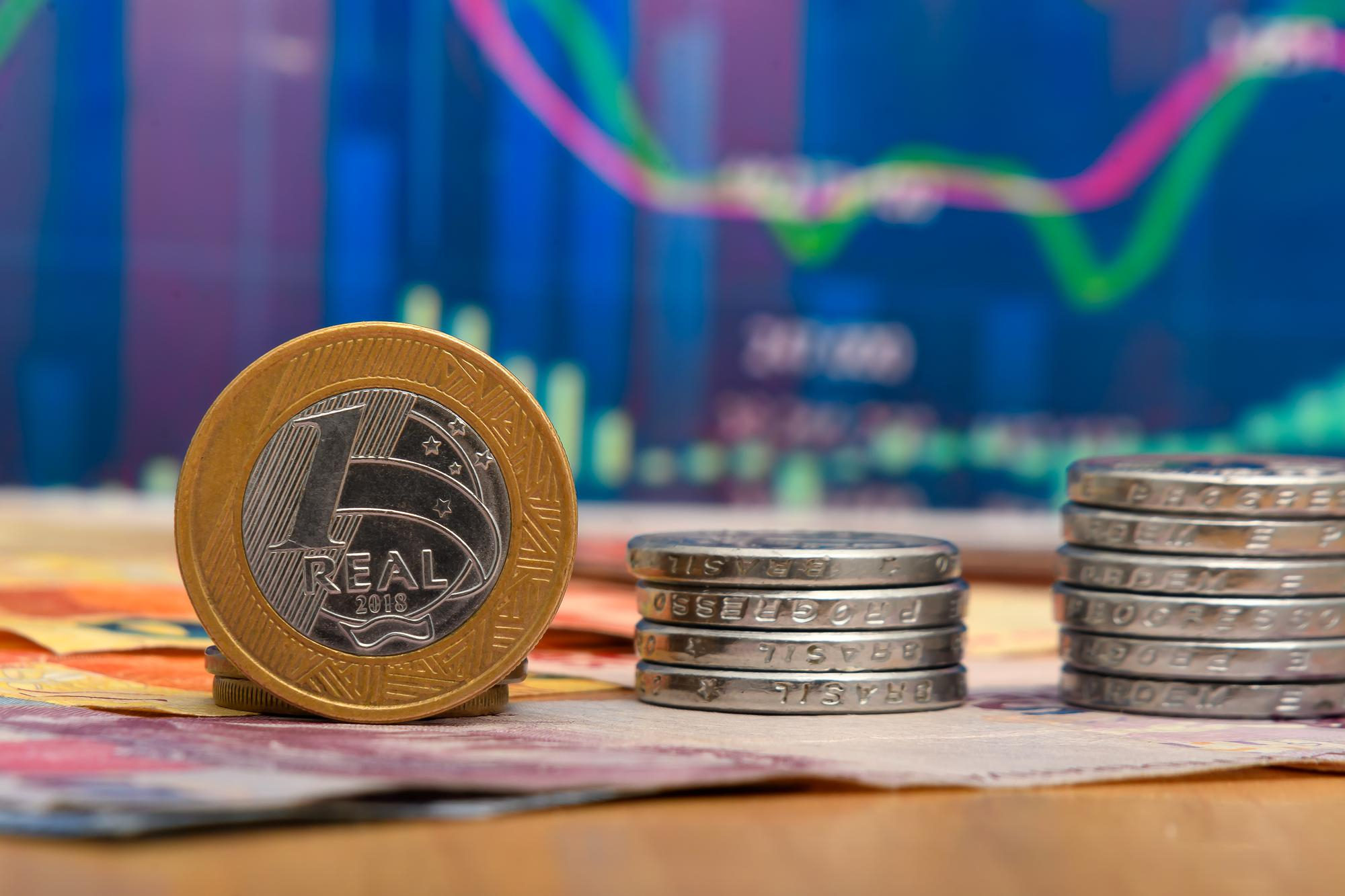 Moeda de 1 real em destaque sobre uma mesa, ao lado de pequenas pilhas de moedas e notas de dinheiro, com um gráfico financeiro colorido desfocado ao fundo. Imagem de capa para o blogpost: Reforma Tributária 2026: Por Que Seu ERP Precisa Estar Preparado Agora