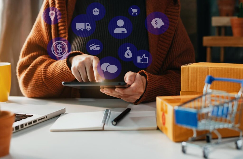 Pessoa usando um tablet em uma mesa com caixas de encomenda, carrinho de compras em miniatura e ícones digitais relacionados a e-commerce e finanças sobrepostos na imagem. Imagem de capa para o blogpost Por que Integrar seu ERP com o Anymarket