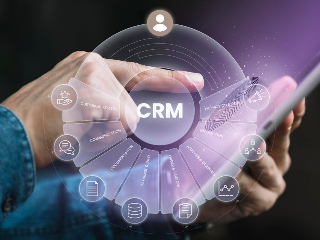 Imagem de capa para o blogpost Mão segurando um smartphone com uma interface holográfica de CRM (Customer Relationship Management), exibindo ícones de comunicação, fidelização de clientes, análise de dados, interações e campanhas promocionais, representando gestão do relacionamento com clientes e uso de tecnologia digital.